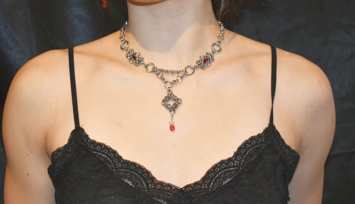 Person wearing a black lace top and an ornate necklace with a red pendant against a dark background