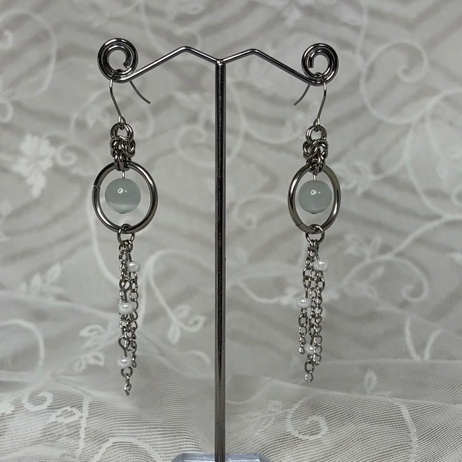 A pair of silver-toned dangle earrings with circular hoops, pale round stones, and dangling chain elements, displayed on a clear stand against a white patterned fabric background.