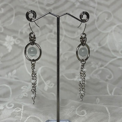 A pair of silver-toned dangle earrings with circular hoops, pale round stones, and dangling chain elements, displayed on a clear stand against a white patterned fabric background.